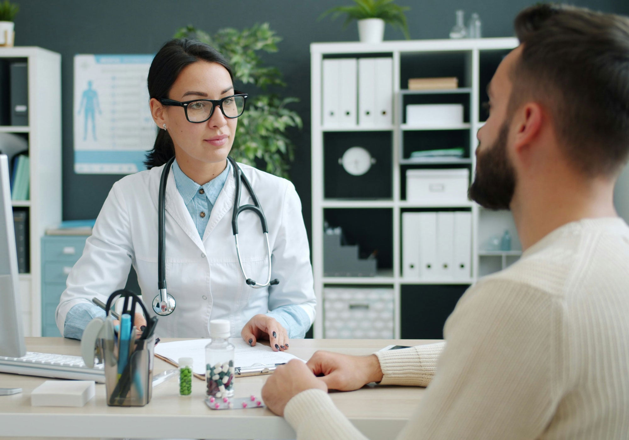 Doctor in a white coat with a stethoscope talking to a patient in an office setting.