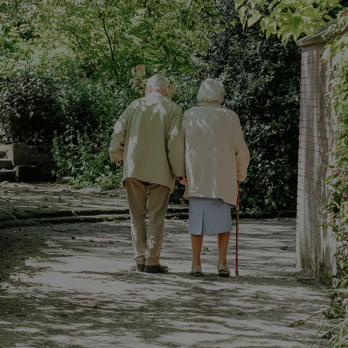 Two elderly people walking together on a path with greenery around.