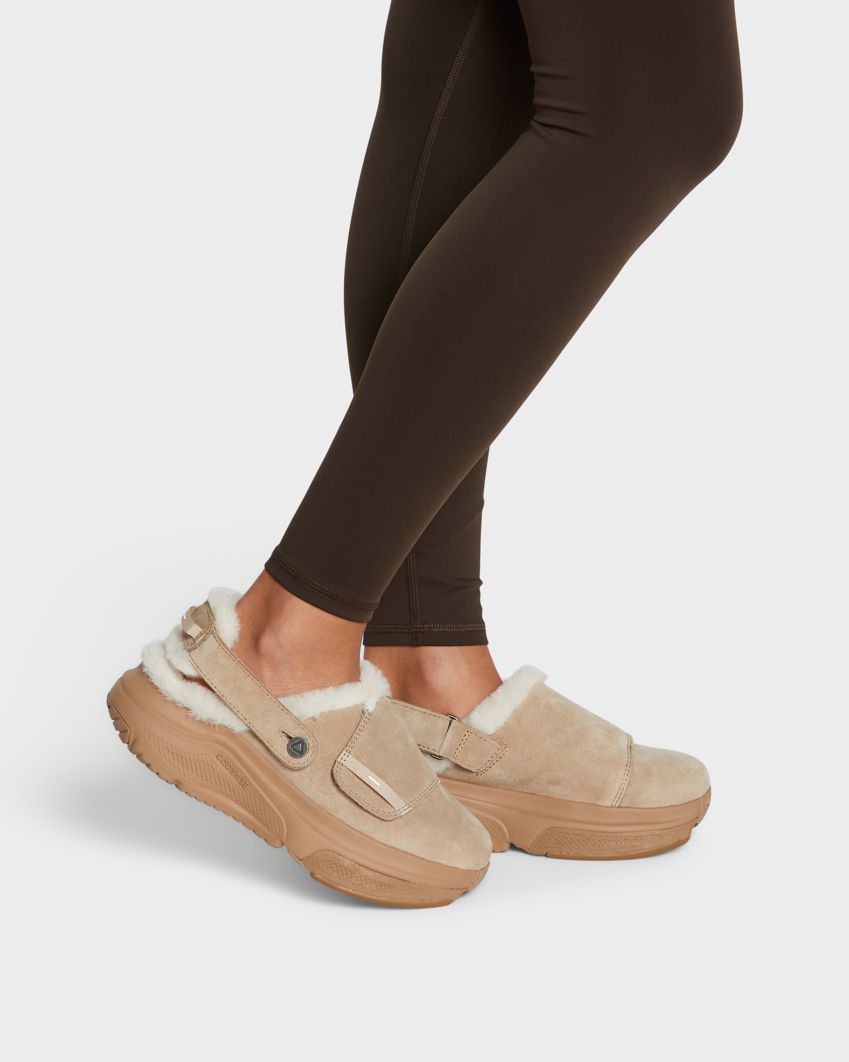 Beige slippers with white fur lining worn with dark leggings on a light gray background