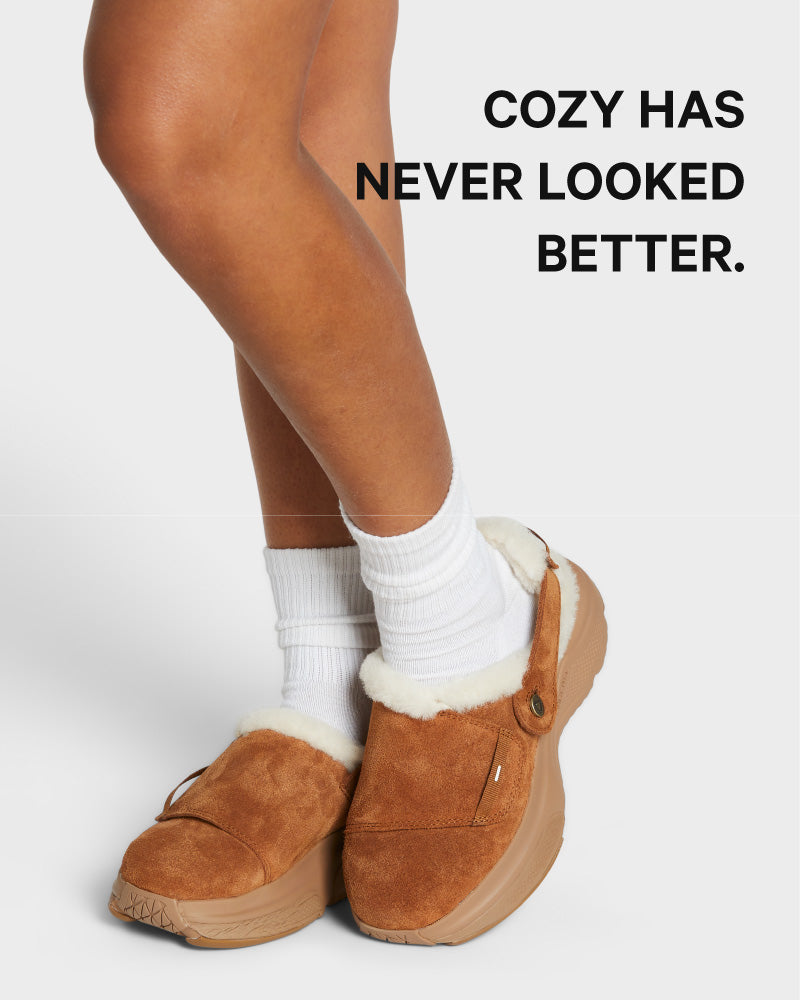 Brown slip-on shoes with white socks on a light gray background, text 'COZY HAS NEVER LOOKED BETTER.'