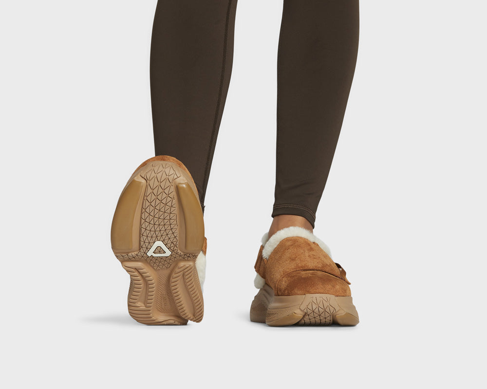 Brown winter slippers with fur lining worn with dark leggings on a white background