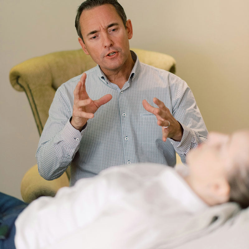 Man in a checkered shirt gesturing with his hands, sitting next to a person lying down.