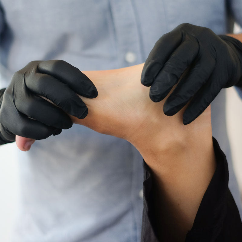 Person wearing black gloves applying a product to their arm against a neutral background