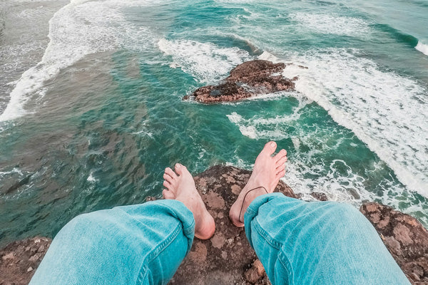 Feet of a person wearing light blue pants overlooking the ocean with waves crashing on rocks.