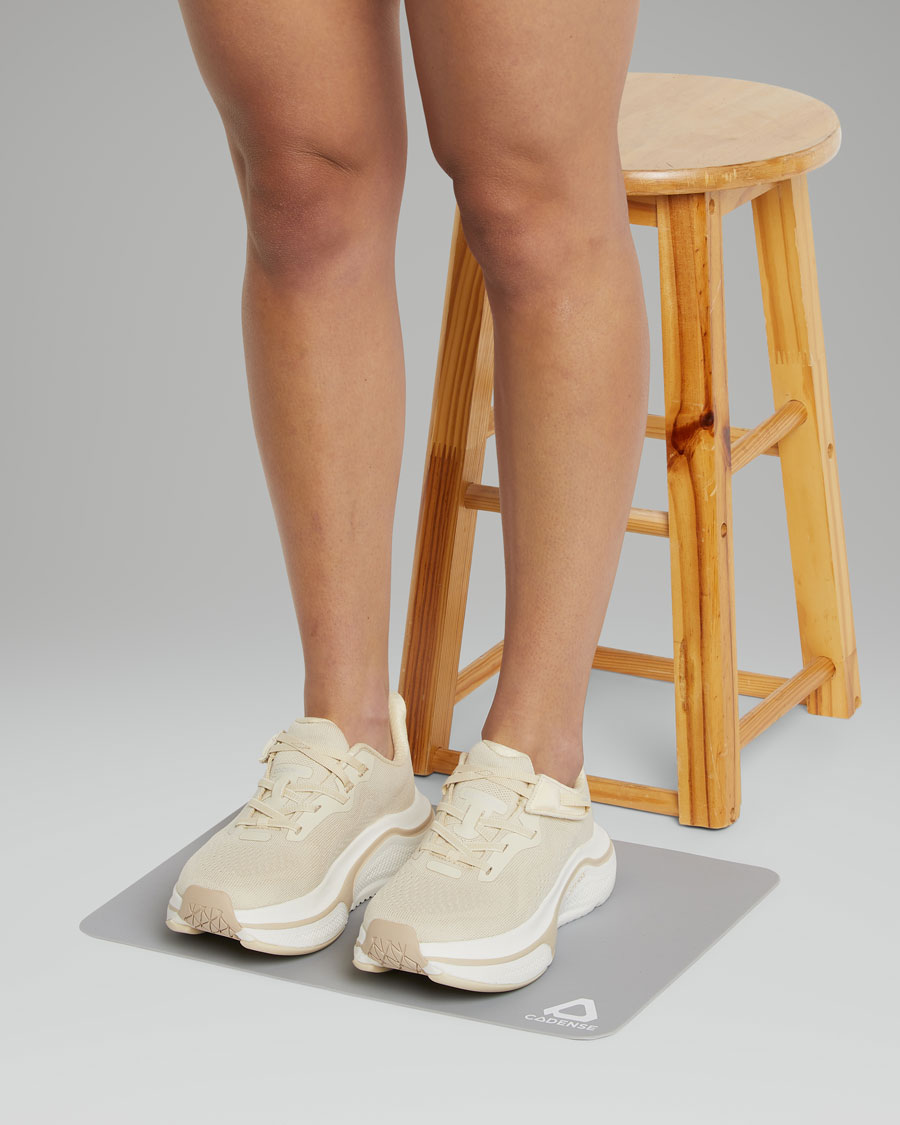 Person wearing beige sneakers on a gray mat with a wooden stool in the background