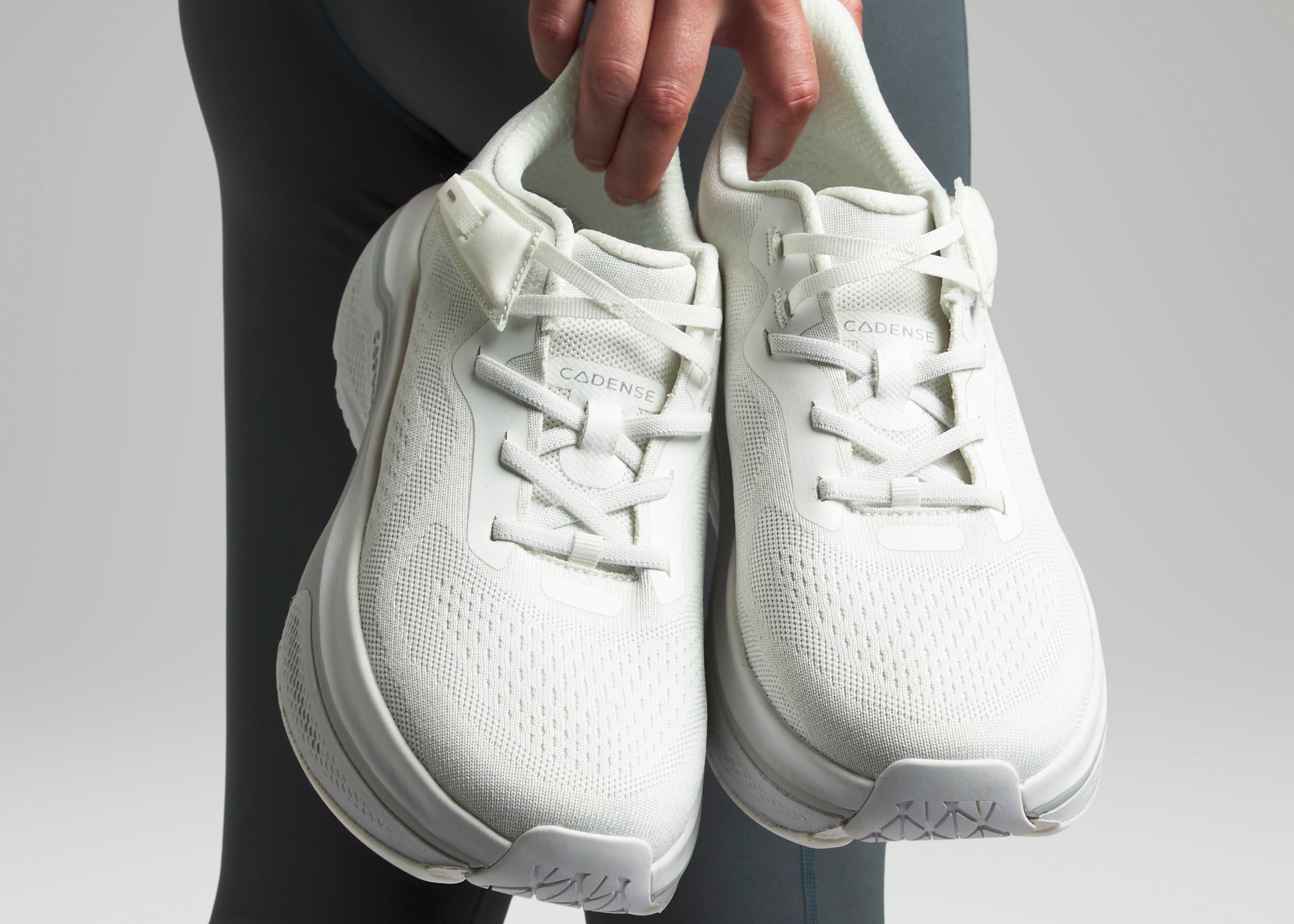 White sneakers held by a person wearing dark leggings on a light gray background