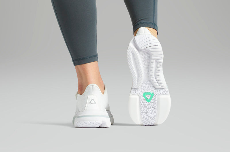 White athletic shoes with a green logo worn by a person in gray leggings on a light gray background