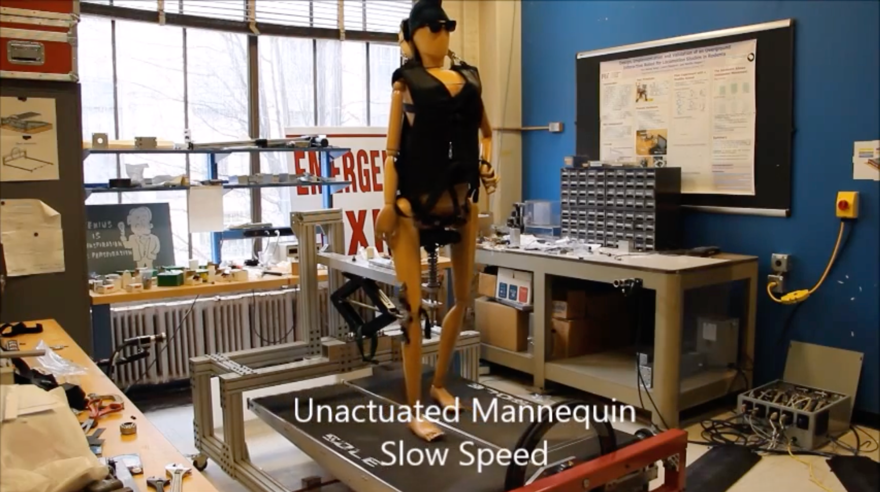 Unactivated mannequin in a laboratory setting with various equipment and a large screen displaying text.